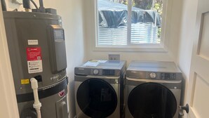 A laundry area with two Samsung washing machines, a water heater, and a window with a view of a covered outdoor area. - Charming 3-bedroom house in Volcano w/laundry & AC, perfect for your getaway! (Volcano)