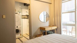 Hotellihuone, jossa on sänky, pyöreä peili, puinen työpöytä, pesukone ja lasinen suihkukaappi.