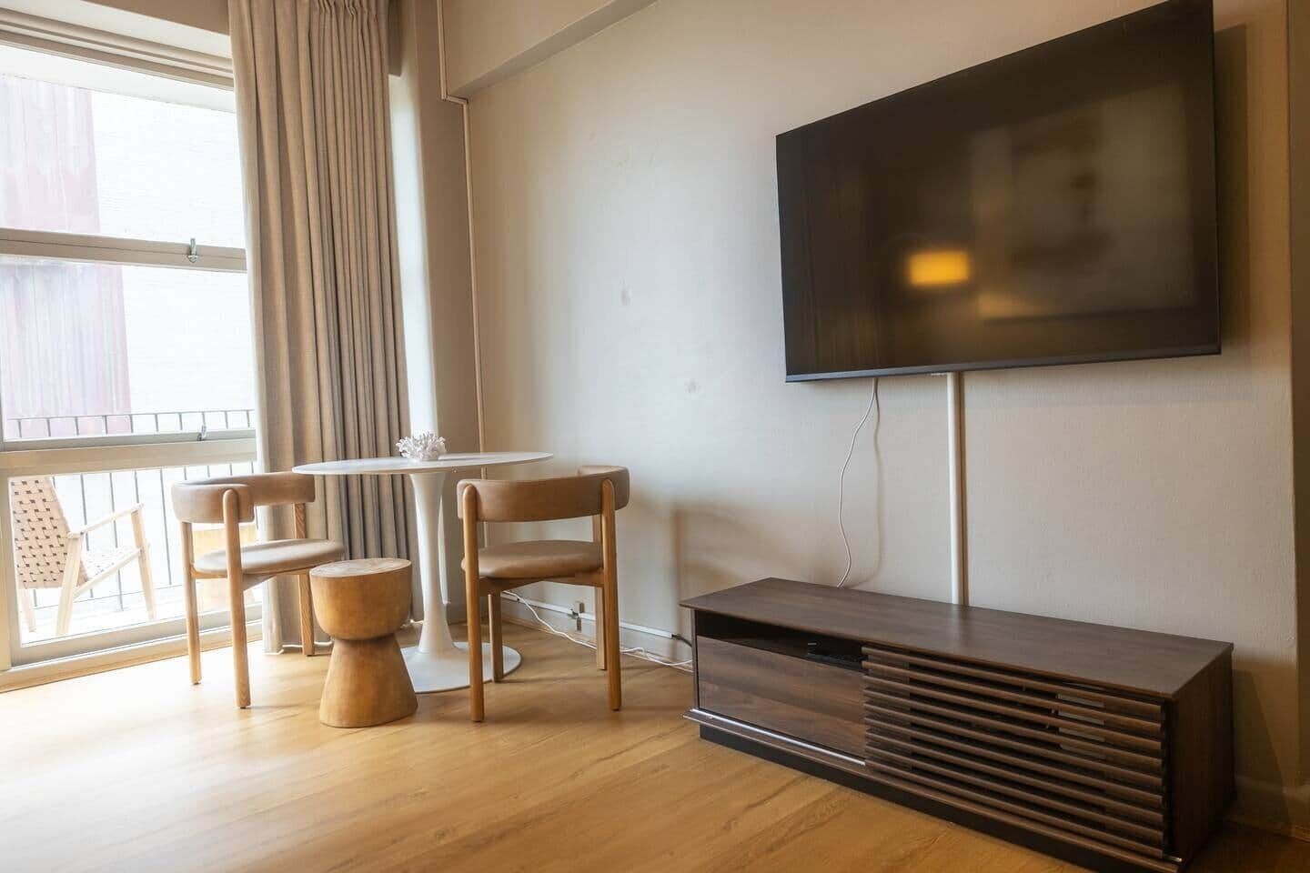 Een moderne woonkamer met een televisie, een houten tv-meubel, een ronde tafel met stoelen en een groot raam met gordijnen.