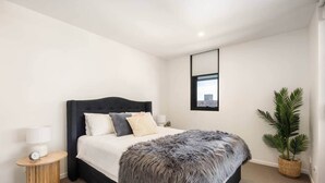 Une chambre à coucher moderne avec une tête de lit sombre, un grand lit, une table de chevet avec une lampe, une petite horloge et une plante en pot.