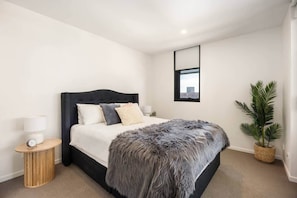 A modern bedroom with a dark headboard, a large bed, a bedside table with a lamp, a small clock, and a potted plant.
