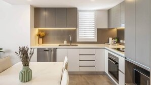 Une cuisine moderne dotée d’un lave-vaisselle intégré, d’une fenêtre avec stores et d’un coin repas agrémenté d’un vase de fleurs séchées.