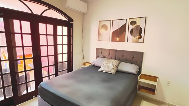 A bedroom with a bed, a chandelier, a window with a view, and artwork on the wall. - Casa com Piscina em Cabeçudas , 80 metros da Praia (Itajaí)