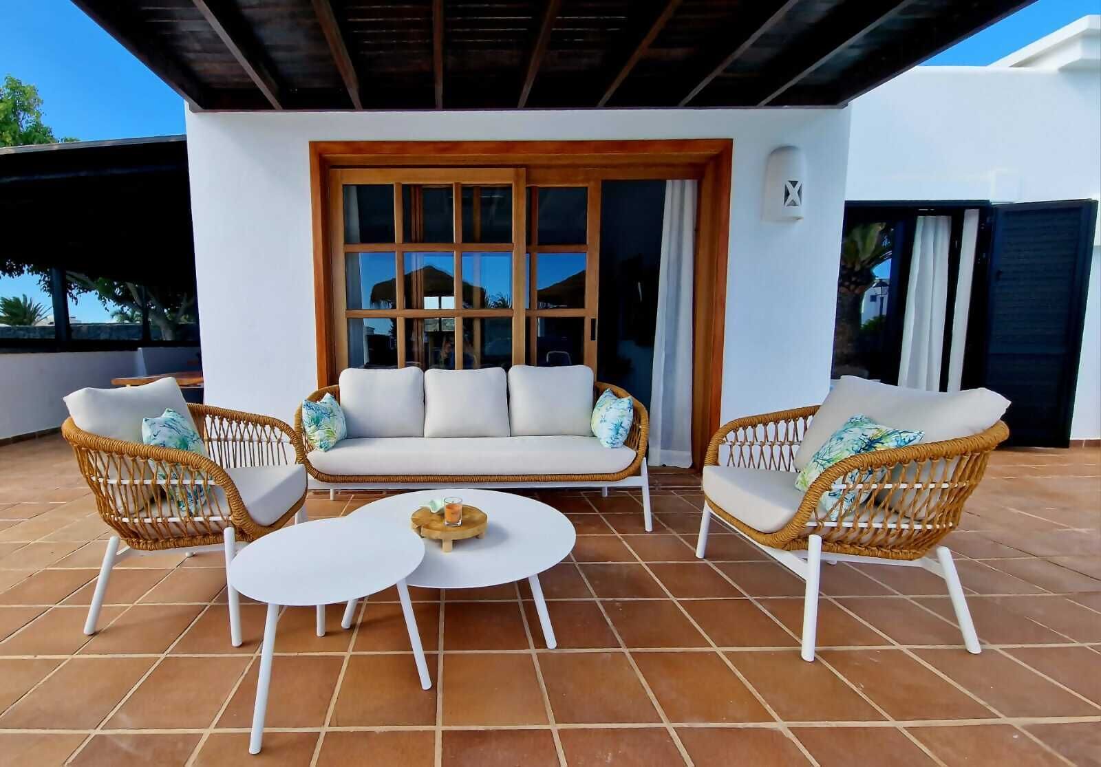 Une terrasse avec un canapé blanc, deux fauteuils en osier, une table ronde et un plateau en bois.