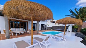 Un espace aménagé au bord de la piscine, avec des chaises longues, un parasol en chaume et une piscine.