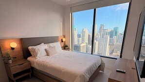 Una camera da letto moderna con un letto grande, comodini, un tavolino e una pianta in vaso, con vista su un orizzonte cittadino attraverso ampie finestre.