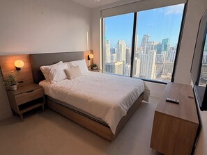 Ein modernes Schlafzimmer mit einem großen Bett, Nachttischen, einem Beistelltisch, einem Topf mit einer Pflanze und Blick auf eine Stadtlandschaft durch große Fenster.