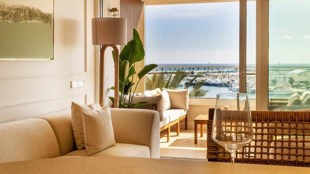 Une chambre d’hôtel moderne dotée d’une grande fenêtre donnant sur le port de plaisance, d’un canapé, d’une table à manger et d’une lampe.