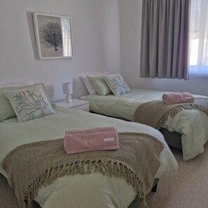 A bedroom with two beds, each with a pillow and a blanket. There is a nightstand with a lamp, a framed picture on the wall, and a window with curtains.