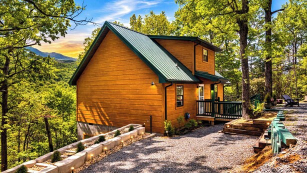 A wooden cabin with a green roof, surrounded by trees and a gravel driveway. - Smoky Ridge Retreat | Hot Tub • Firepit • Views (Gatlinburg)