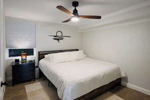 Une chambre à coucher comprenant un lit, une table de chevet, une lampe, un ventilateur de plafond et une fenêtre avec des stores.
