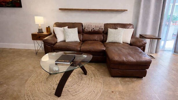 Ein braunes, ledernes Ecksofa mit einem Glastisch, einem kleinen Beistelltisch mit Lampe und einem Teppich auf Fliesenboden.