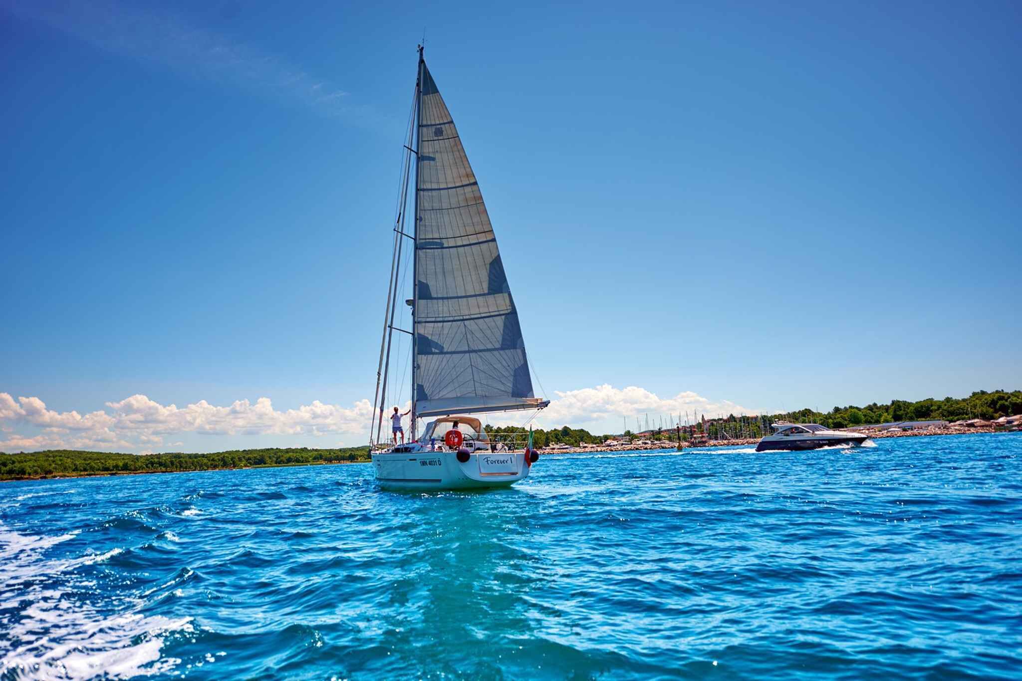 A sailboat with a person standing at the stern, another person on the deck, and a coastline with buildings in the background.