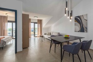 Ein modernes Wohnzimmer mit Sofa, Esstisch und Stühlen, einem großen Fenster mit Vorhängen und einer Deckenleuchte.