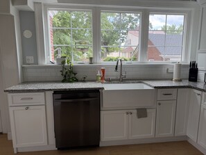 A kitchen with white cabinetry, a dishwasher, a sink, and a window overlooking a residential area. - Lux Colonial Home: Hot tub, Game Room, Home Theatre, Karaoke (Framingham)