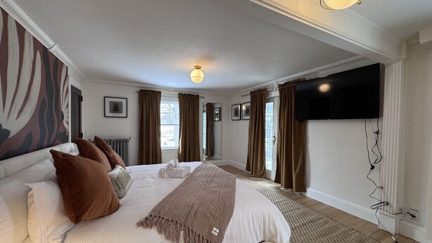 Un dormitorio con una cama grande, un televisor de pantalla plana montado en la pared y un ventanal con cortinas.