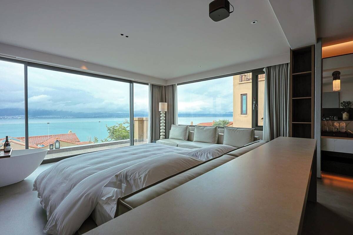 A modern hotel room with a large bed, a bedside table, a lamp, and a view of the sea.