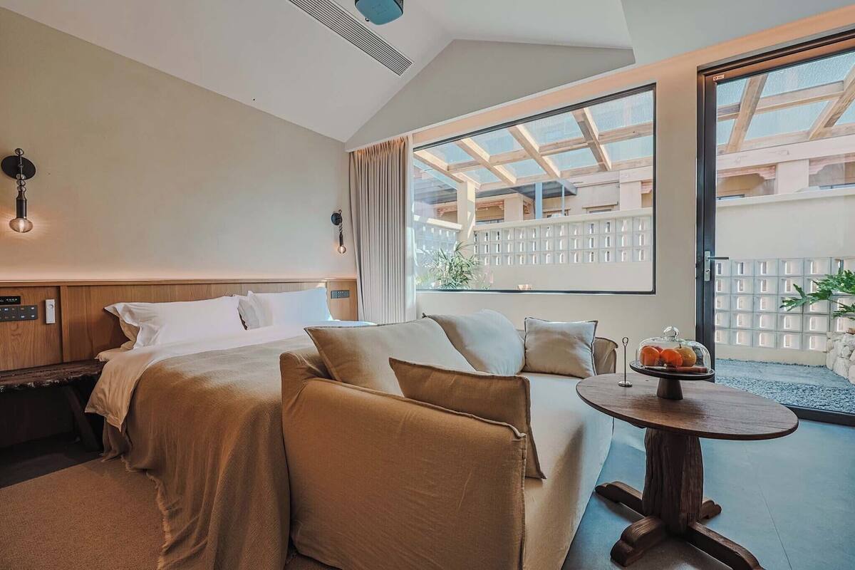 A hotel room with a bed, a sofa, a small table with fruit, a window with a view of a building, and a ceiling fan.
