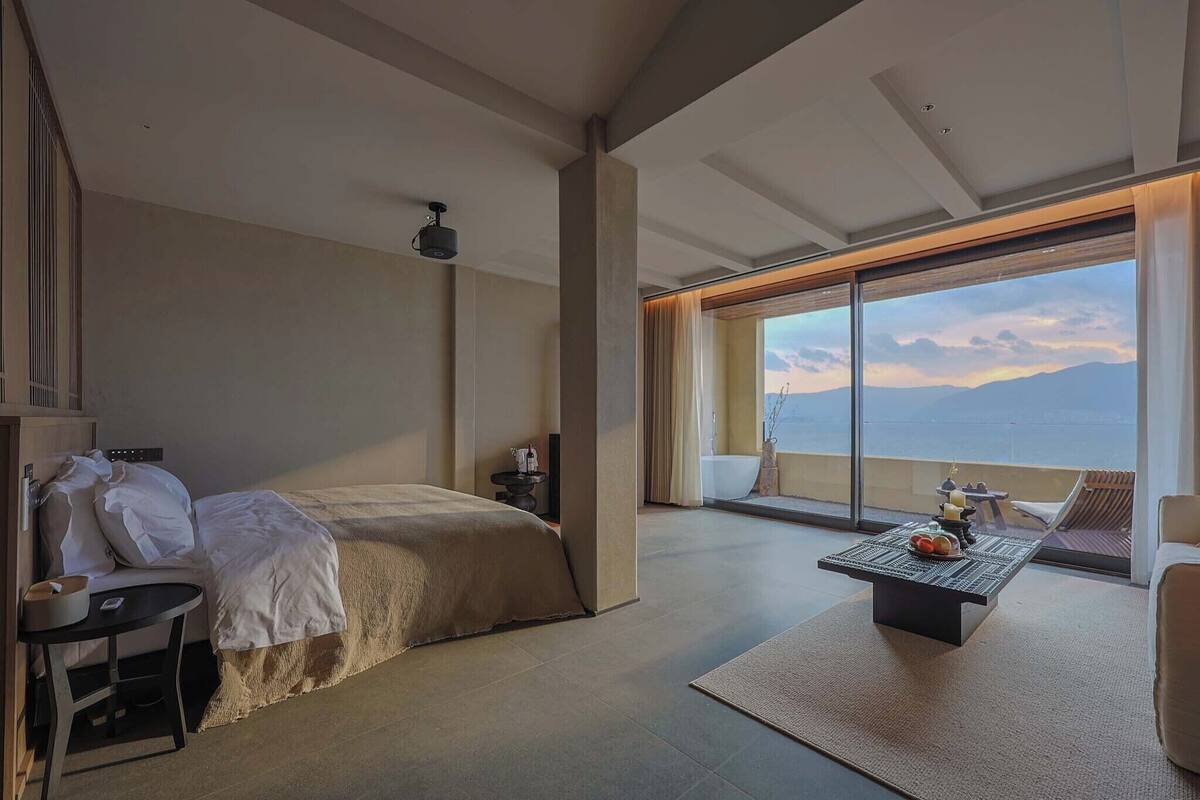A modern hotel room with a bed, a small table, a chair, and a large window offering a view of the mountains.