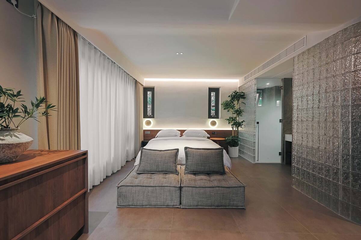 A modern hotel room with a large bed, bedside tables, a wooden dresser, and a potted plant.