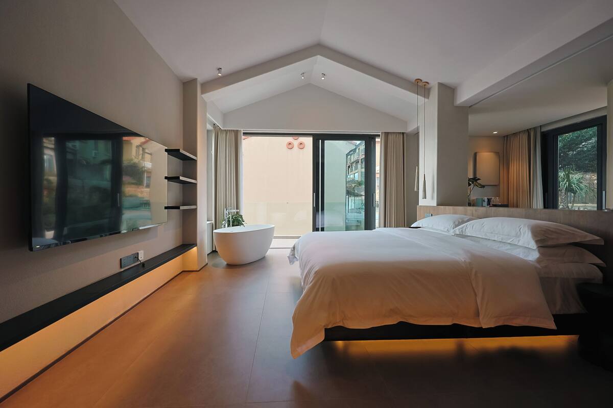 A modern bedroom with a large bed, a flat-screen TV mounted on the wall, a built-in bathtub, and a view through a glass door.