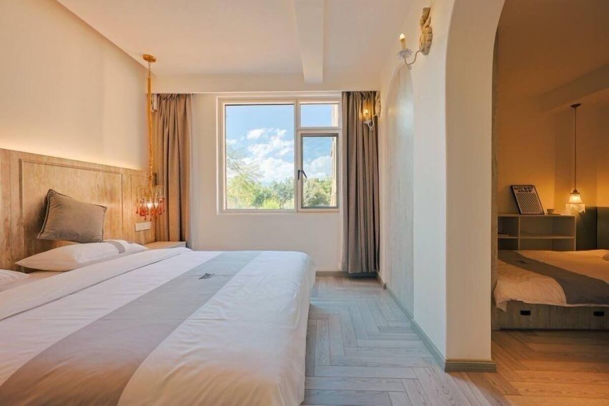A hotel room with a large bed, a window with curtains, a wooden headboard, and a small seating area with a lamp.