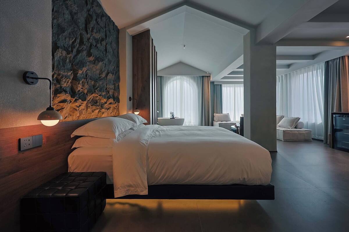A modern hotel room with a large bed, a bench, a wall-mounted lamp, and a view of another room.