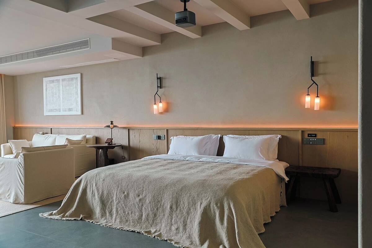 A hotel room with a large bed, a sofa, a small table, and wall-mounted lighting.