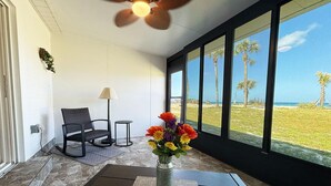 A room with a ceiling fan, a chair, a table, a vase with flowers, and a view of the beach. - Venice Villas San Marco - Oceanfront - Beach (Venice)