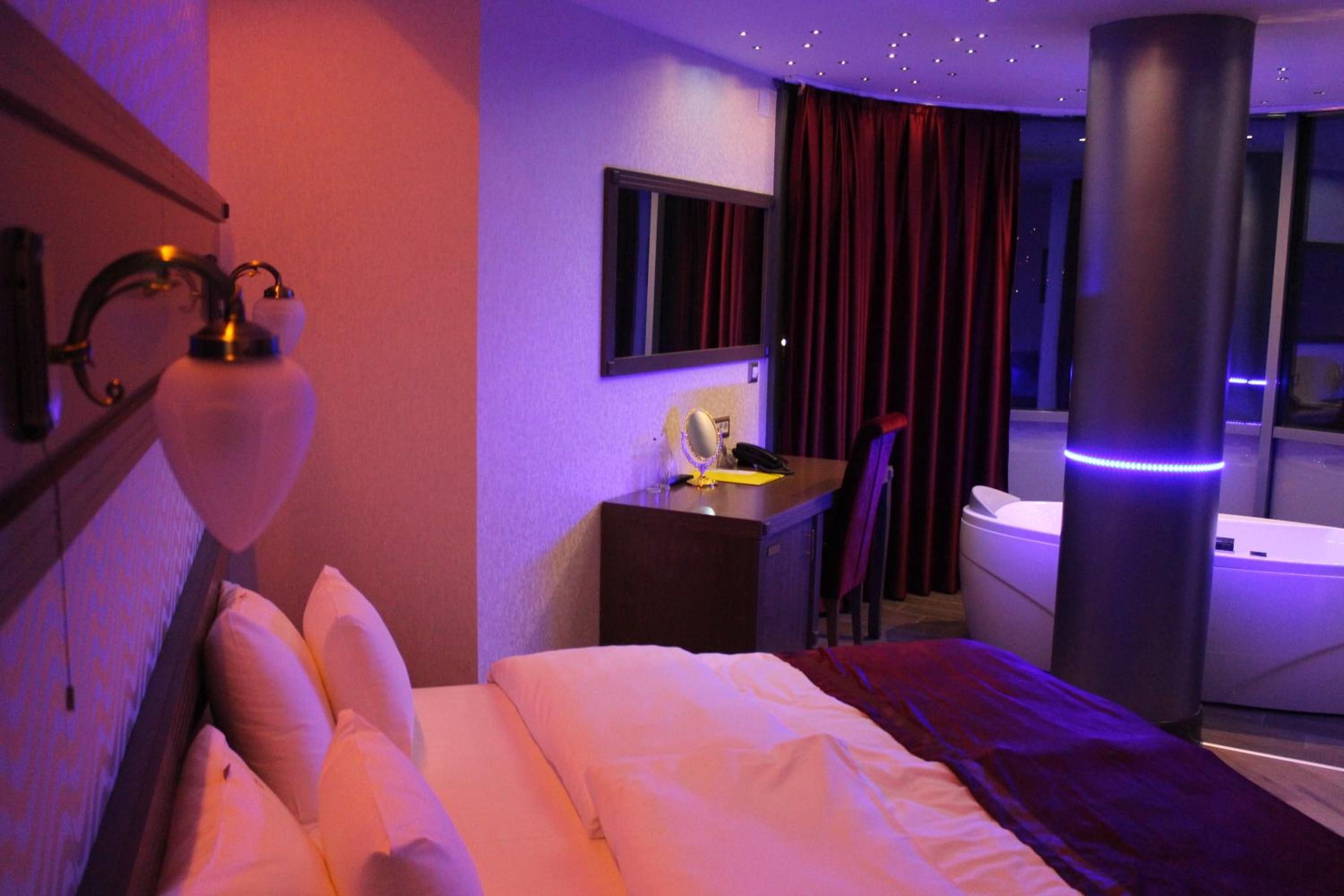 Quarto de hotel com cama, escrivaninha, cadeira, espelho, janela com cortinas e banheira.