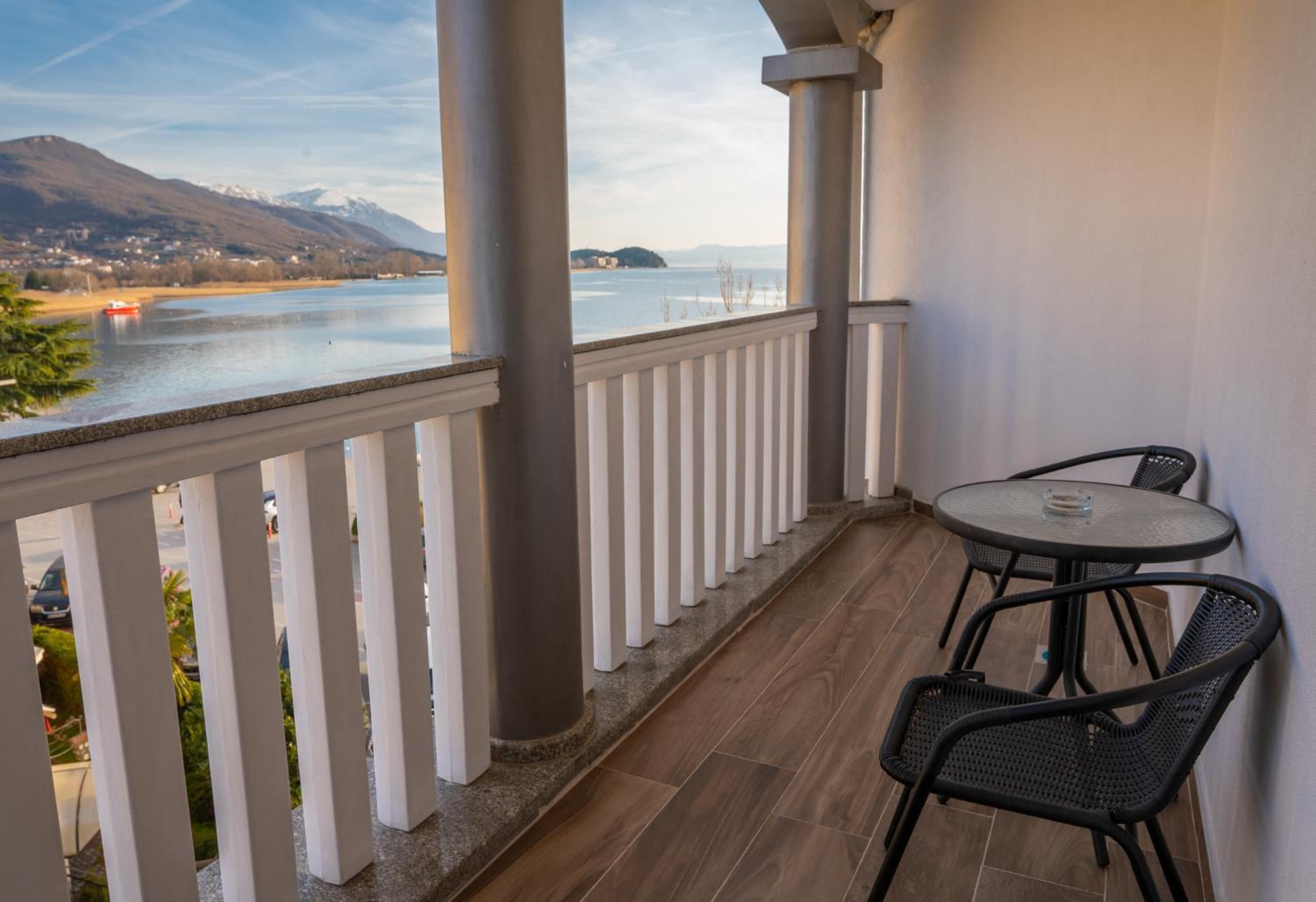 A balcony with a view of a lake and mountains, a round table with chairs, and a white railing.