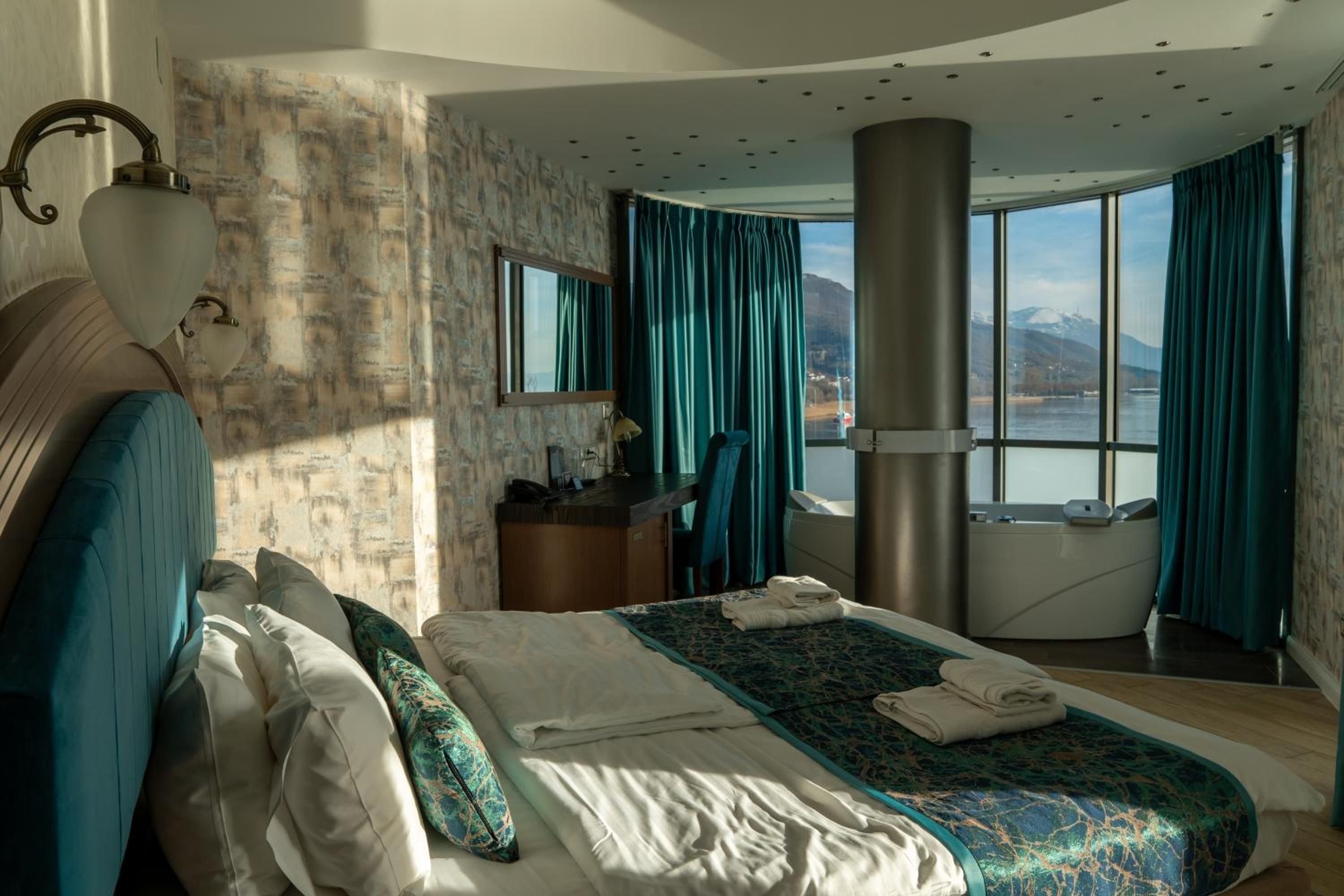 Een hotelkamer met een groot bed, een bureau, een stoel, een spiegel en uitzicht op een meer en bergen.