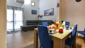 A living room with a sofa, a dining table with chairs, a clock, and a window with curtains. - 2 bedroom lovely home in Castellammare del Golf (Castellammare del Golf)