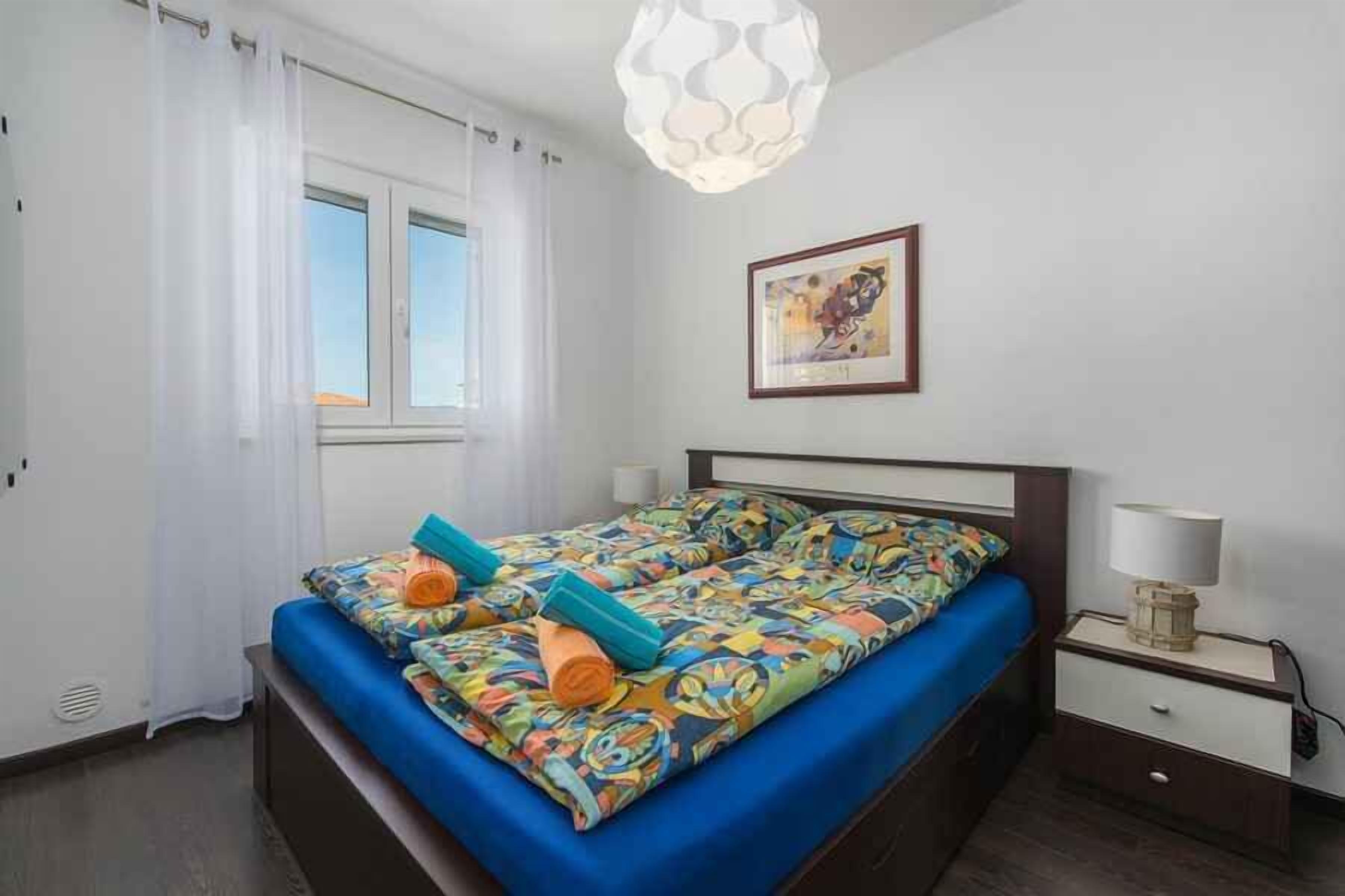 Ein Schlafzimmer mit einem Bett, einem Nachttisch, einer Lampe, einem Fenster mit Vorhängen und einem Wandbild.