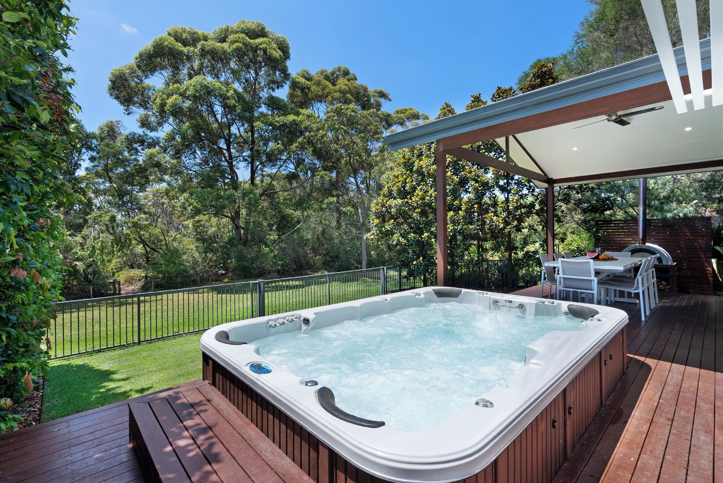 A hot tub on a wooden deck with a covered patio area, surrounded by trees and a fenced yard.