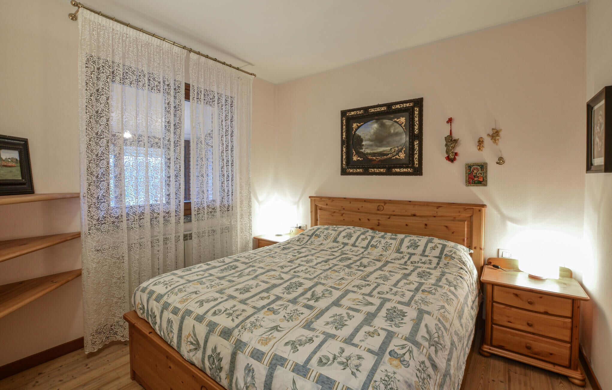 Quarto com cama de madeira, mesa de cabeceira, janela com cortinas de renda e um quadro pendurado na parede.