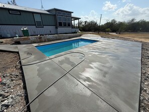 Une piscine avec une terrasse en béton, une maison en arrière-plan et un ciel dégagé.