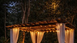 A wooden gazebo with draped curtains and string lights, surrounded by potted plants and yoga mats.