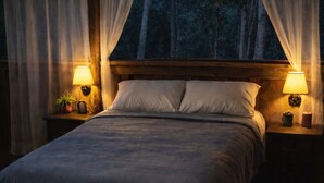 A bed with a wooden headboard, two bedside lamps, and a view of trees through a window with sheer curtains.