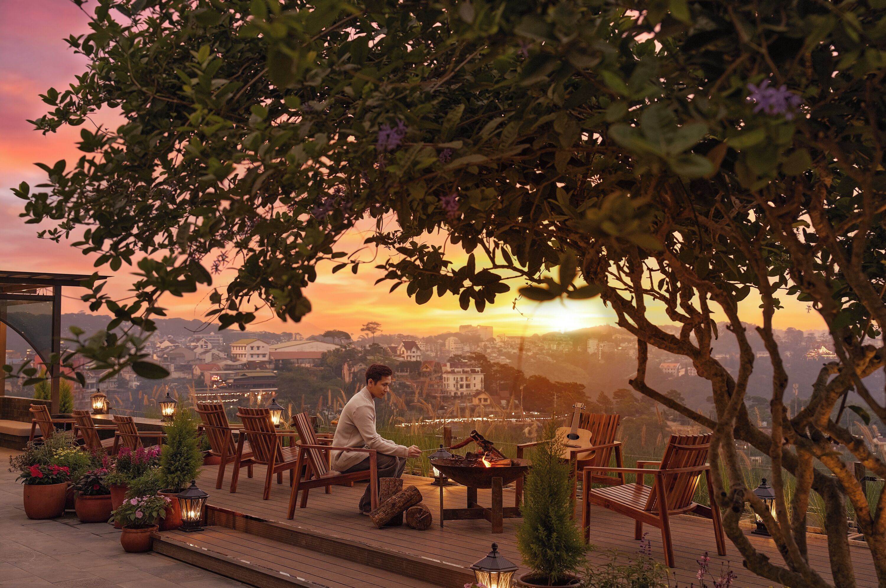 A man sitting by a fire pit on a deck with chairs and tables, overlooking a cityscape at sunset.