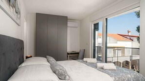 A bedroom with a bed, a desk, and a chair, a wardrobe, and a large window with a view of buildings. - Holiday Apartment Sabbia 6+0 (Srima)