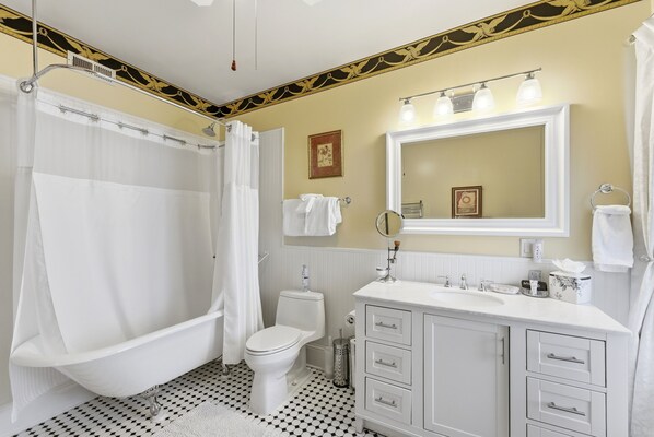 Une salle de bain comprenant une baignoire sur pieds, des toilettes, un meuble-lavabo avec des tiroirs, un miroir et un rideau de douche.