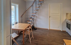 Una habitación con una mesa de madera, sillas, una escalera, una puerta blanca y una zona de cocina.