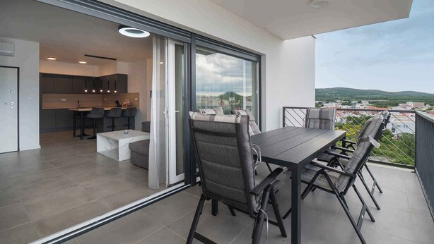 Un balcone moderno con tavolo e sedie, un'area cucina visibile tramite porte in vetro e vista sul paesaggio circostante.