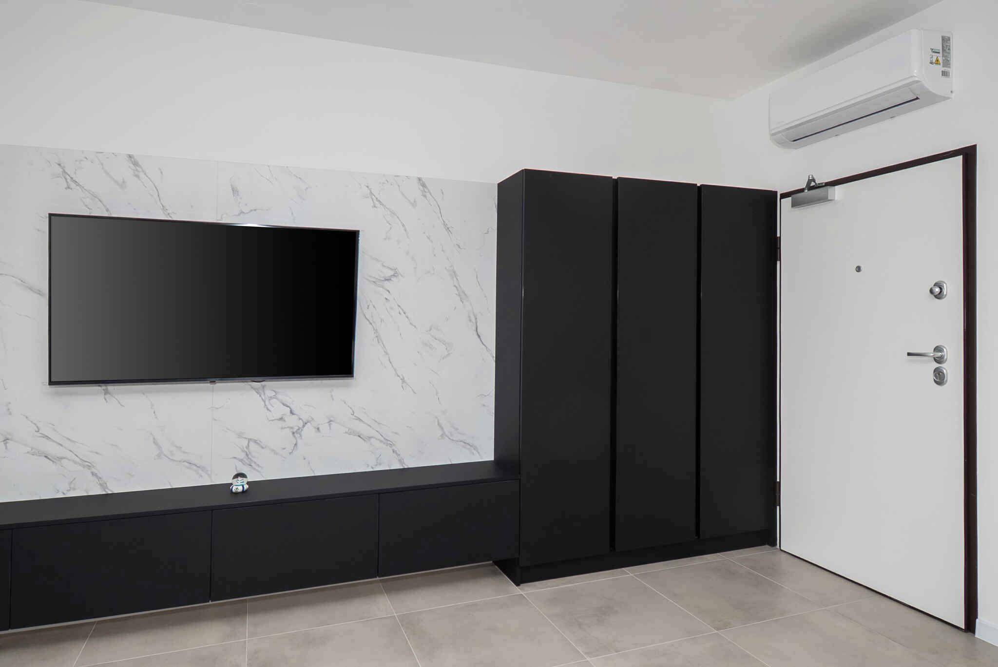 Une chambre moderne avec un téléviseur à écran plat fixé sur un mur en marbre, un meuble de rangement noir et une porte blanche dotée d’une poignée.