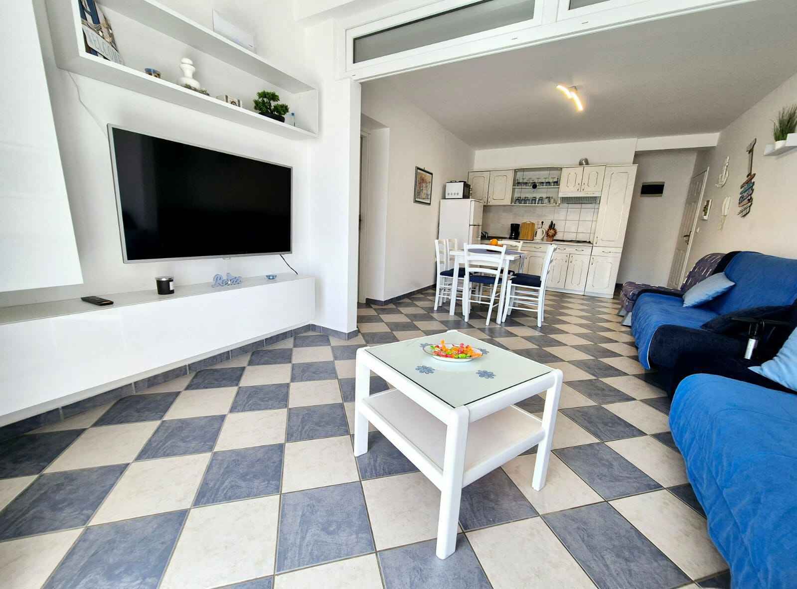 A modern living room with a flat-screen TV, a dining area with a table and chairs, a kitchen with cabinets, and a blue sofa.