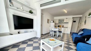 Un salon moderne doté d’un téléviseur à écran plat, d’un coin repas avec une table et des chaises, d’une cuisine équipée de placards et d’un canapé bleu.