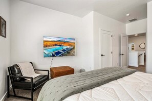 Une chambre à coucher comprenant un lit, une chaise, un téléviseur fixé au mur et une porte donnant sur une autre pièce.