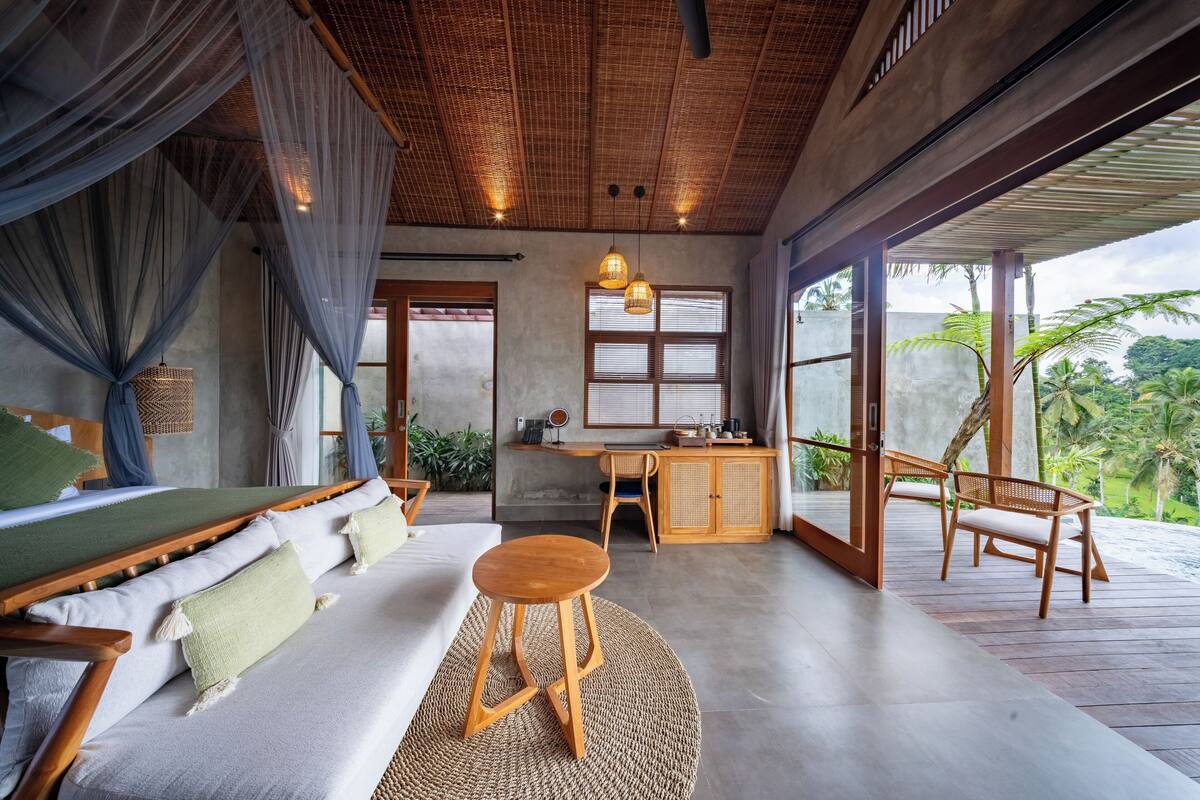 Une pièce spacieuse avec un plafond en bois, une grande fenêtre, un bureau en bois, un canapé et un petit tabouret.