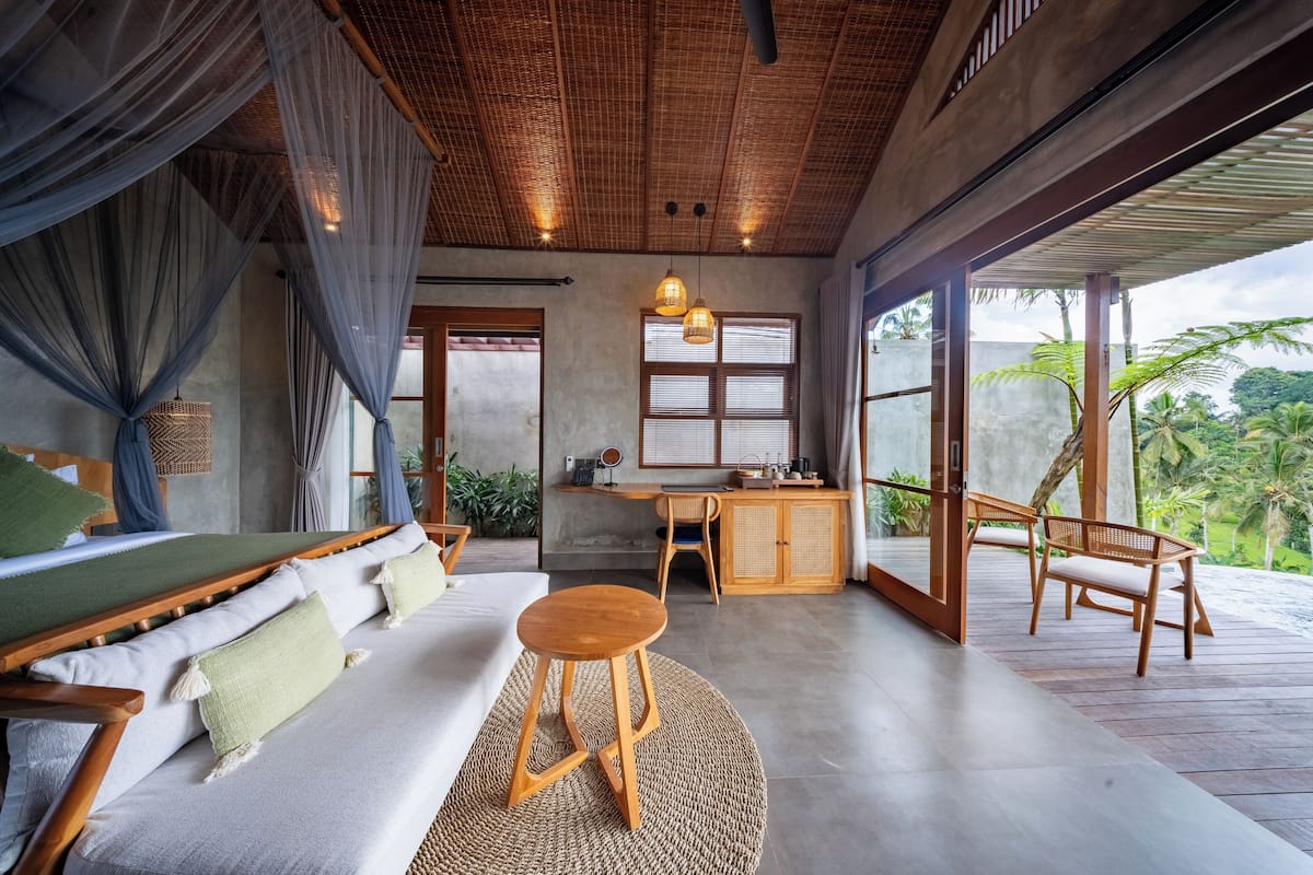 A spacious room with a wooden ceiling, a large window, a wooden desk, a sofa, and a small stool.